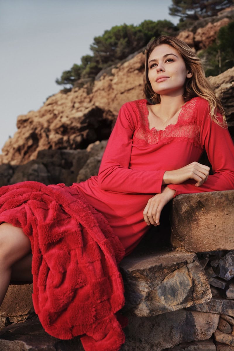 Eine Frau lächelt, trägt ein rotes Kleid mit Spitzen, sitzt auf Felsen vor einer felsigen Landschaft.